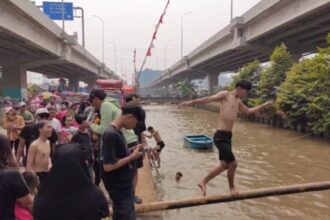 Peserta lomba titian bambu menjaga keseimbangan berjalan di atas pohon pinang dilumur oli agar tidak tercebur di aliran Kalimalang dalam lomba diadakan warga RW 04, Cipinang Melayu, Makasar, Jakarta Timur, pada Sabtu (24/8/2024) siang. Foto: Joesvicar Iqbal/ipol.id