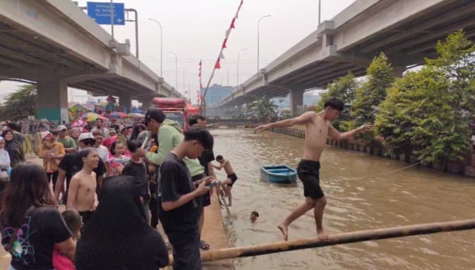 Peserta lomba titian bambu menjaga keseimbangan berjalan di atas pohon pinang dilumur oli agar tidak tercebur di aliran Kalimalang dalam lomba diadakan warga RW 04, Cipinang Melayu, Makasar, Jakarta Timur, pada Sabtu (24/8/2024) siang. Foto: Joesvicar Iqbal/ipol.id