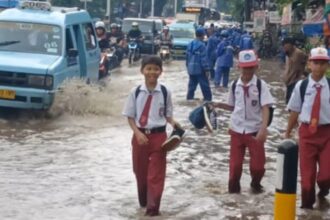 Suasana tiga siswa Sekolah Dasar (SD) berjalan kaki menerabas genangan di Jalan Raya Bogor, Kramat Jati, Jakarta Timur, mengarah traffick light Pusat Grosir Cililitan, Senin (12/1/2026) siang. Foto: Joesvicar Iqbal/ipol.id